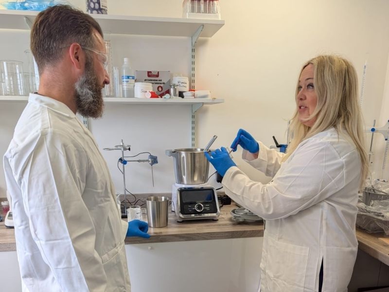 Two formulation scientists in white lab coats and blue gloves working at a bench with a stainless-steel mixing vessel